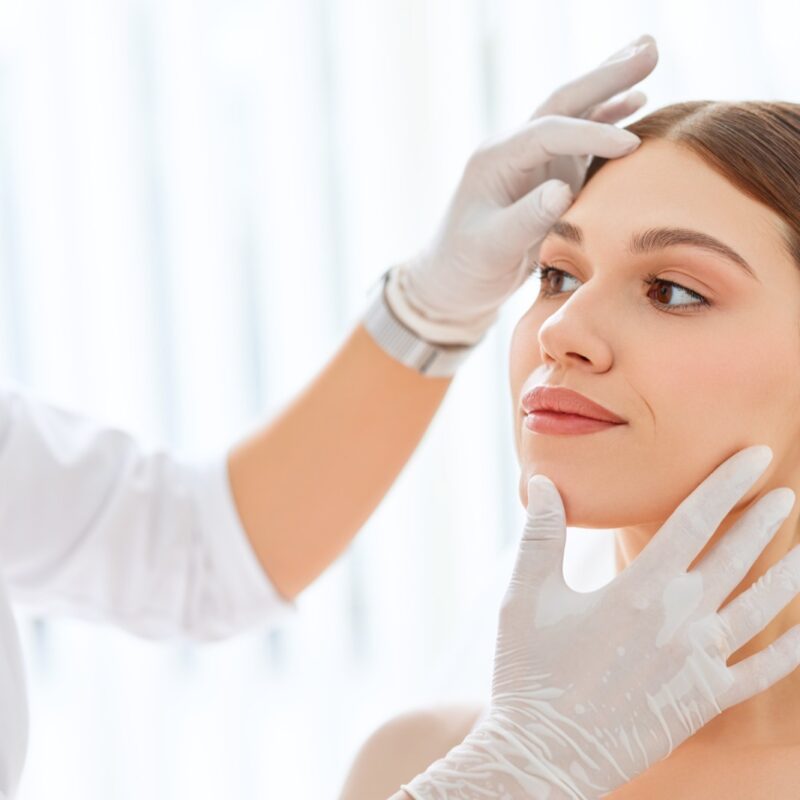 Crop cosmetologist in white gloves examining face skin of young female client before beauty procedure in salon